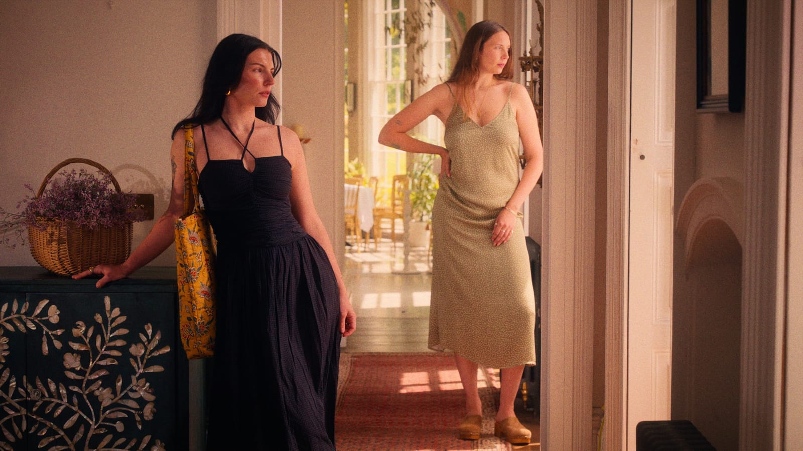 Two women standing in a sunlit doorway, one leaning on a painted cabinet, both looking off to the left